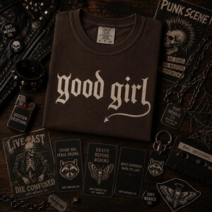 Espresso brown Comfort Colors 1717 “good girl” gothic graphic t-shirt folded on dark walnut wood surface with moody cinematic lighting. Punk-inspired alternative fashion flat lay featuring chains, stickers, leather textures, and Soft Menace Co. branding details. Vintage garment dyed cotton t-shirt with distressed cream blackletter typography for women who love edgy streetwear and gothic apparel.