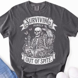 Pepper Comfort Colors 1717 skeleton shirt flat lay with jeans and white sneakers. Gothic illustration with coffin, roses, butterflies. Dark academia shirt — Surviving Purely Out Of Spite. DTG printed.