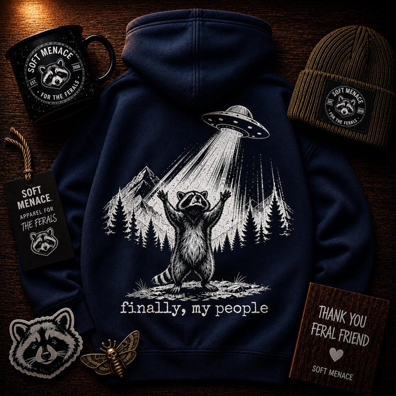 Op de afbeelding: Marineblauwe hoodie met een wasbeer-afbeelding die door een UFO wordt opgetild, de tekst luidt "finally, my people". Ook afgebeeld zijn een zwarte mok, een groene muts, een label, een patch en een houten bord, allemaal met het "Soft Menace" logo.