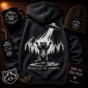 Op de afbeelding: Donkergrijze hoodie met een wasbeer-afbeelding die door een UFO wordt opgetild, met de tekst "finally, my people". Ook afgebeeld zijn een zwarte mok, een groene muts, patches en een houten bord. De items zijn voorzien van "Soft Menace".