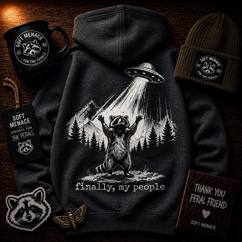 Op de afbeelding: Donkergrijze hoodie met een wasbeer-afbeelding die door een UFO wordt opgetild, met de tekst "finally, my people". Ook afgebeeld zijn een zwarte mok, een groene muts, patches en een houten bord. De items zijn voorzien van "Soft Menace".
