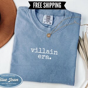 Blue Jean Comfort Colors 1717 tee with villain era printed in white Special Elite typewriter font. Folded flat lay styled with brown cowboy hat, gold chain necklace, and dried palm leaf on white background. Dark humor evil queen graphic tee.