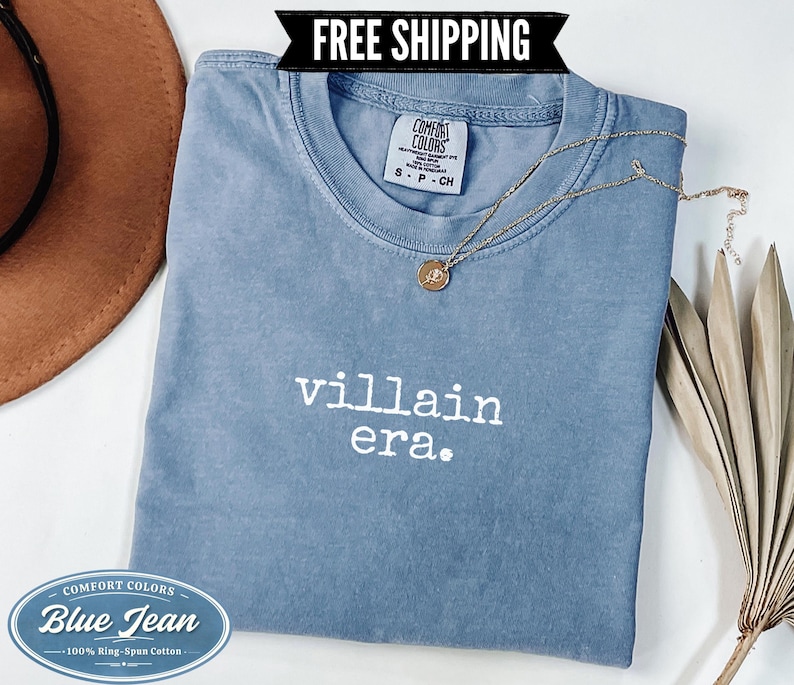 Blue Jean Comfort Colors 1717 tee with villain era printed in white Special Elite typewriter font. Folded flat lay styled with brown cowboy hat, gold chain necklace, and dried palm leaf on white background. Dark humor evil queen graphic tee.
