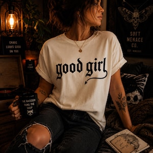 Woman wearing ivory Comfort Colors 1717 “good girl” gothic graphic t-shirt with relaxed fit and dark feminine styling. Moody cinematic lighting, ripped black jeans, layered jewelry, and warm alternative aesthetic create premium editorial streetwear scene. Soft Menace Co. vintage garment dyed graphic tee with distressed gothic typography for women who love punk-inspired casual fashion.