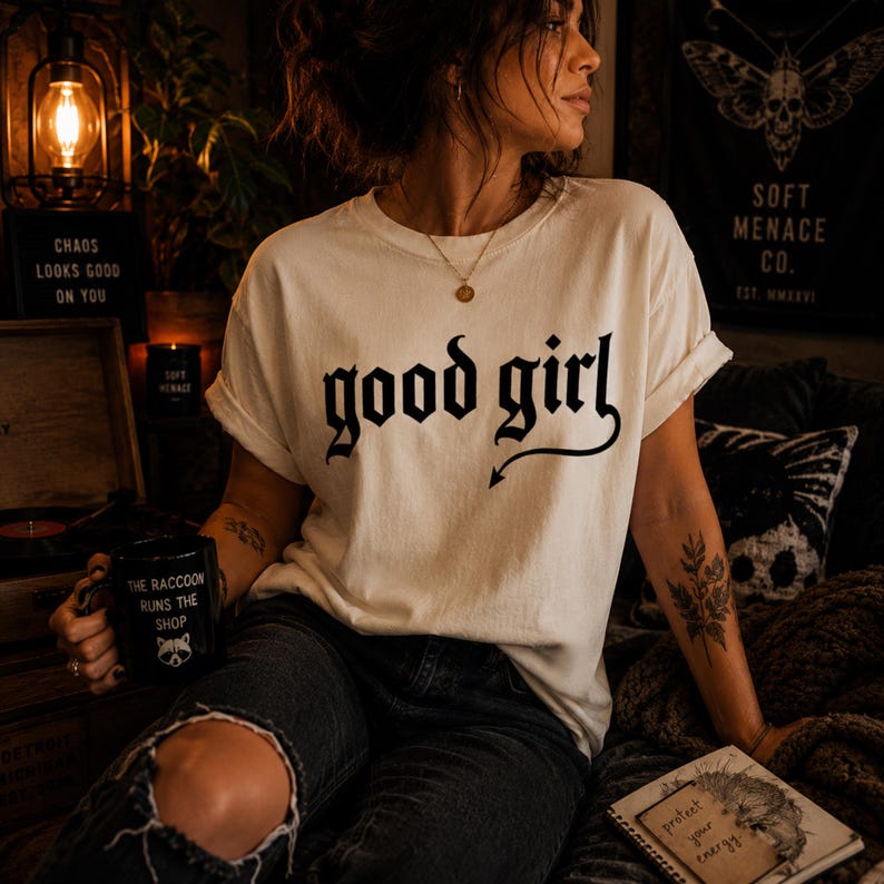 Woman wearing ivory Comfort Colors 1717 “good girl” gothic graphic t-shirt with relaxed fit and dark feminine styling. Moody cinematic lighting, ripped black jeans, layered jewelry, and warm alternative aesthetic create premium editorial streetwear scene. Soft Menace Co. vintage garment dyed graphic tee with distressed gothic typography for women who love punk-inspired casual fashion.