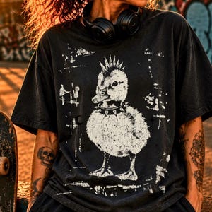 Puede incluir: Una camiseta negra de manga corta con un estampado gráfico blanco de un patito con cresta mohicana y un collar de pinchos. El diseño incluye marcas blancas texturizadas y desgastadas que rodean al patito en la parte frontal de la prenda.