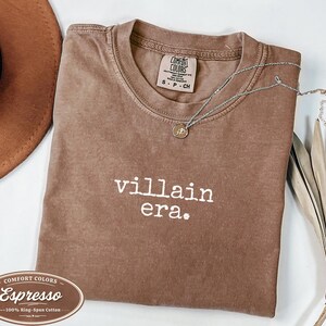 Espresso Comfort Colors 1717 tee with villain era in white Special Elite typewriter font. Folded flat lay with brown cowboy hat, gold necklace, and dried palm leaf. Rich brown garment dyed villain era graphic tee.