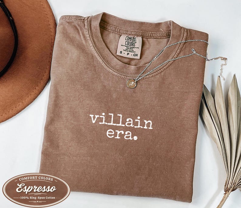 Espresso Comfort Colors 1717 tee with villain era in white Special Elite typewriter font. Folded flat lay with brown cowboy hat, gold necklace, and dried palm leaf. Rich brown garment dyed villain era graphic tee.