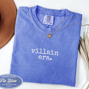 Flo Blue Comfort Colors 1717 tee with villain era in white Special Elite typewriter font. Folded flat lay with brown cowboy hat, gold necklace, and dried palm leaf. Trendy dark academia graphic tee.