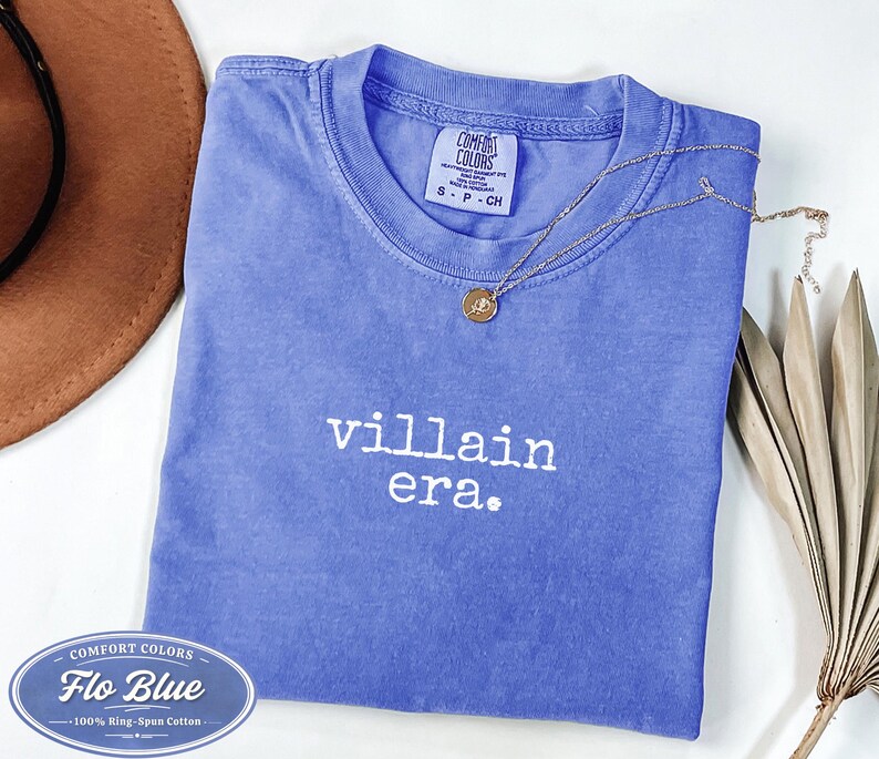 Flo Blue Comfort Colors 1717 tee with villain era in white Special Elite typewriter font. Folded flat lay with brown cowboy hat, gold necklace, and dried palm leaf. Trendy dark academia graphic tee.