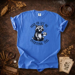 May include: Blue t-shirt featuring a raccoon holding a coffee mug. The raccoon illustration is black and white with large eyes. Text on the shirt reads "Hold on let me overthink this". The coffee mug says "Soft Menace".