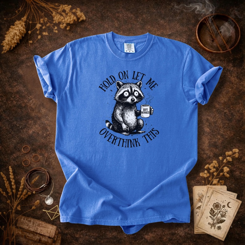 May include: Blue t-shirt featuring a raccoon holding a coffee mug. The raccoon illustration is black and white with large eyes. Text on the shirt reads "Hold on let me overthink this". The coffee mug says "Soft Menace".