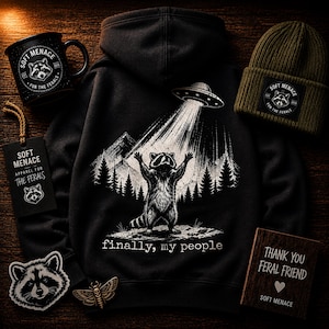 Op de afbeelding: Een zwarte hoodie, mok en beanie met een wasbeerontwerp dat door een UFO wordt opgetild, met de tekst "finally, my people". Bevat ook een label, patch en houten bord met de woorden "Soft Menace" en "Thank You Feral Friend".