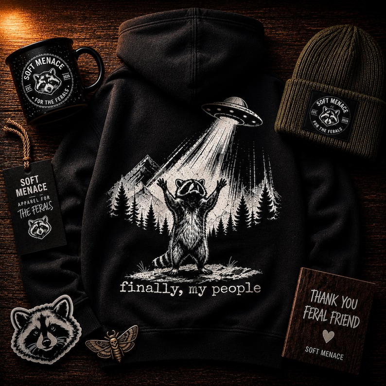 Op de afbeelding: Een zwarte hoodie, mok en beanie met een wasbeerontwerp dat door een UFO wordt opgetild, met de tekst "finally, my people". Bevat ook een label, patch en houten bord met de woorden "Soft Menace" en "Thank You Feral Friend".