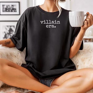 May include: Black t-shirt with the text "villain era." in white. The shirt is worn by a person holding a white mug. The person is sitting on a fluffy, white surface.