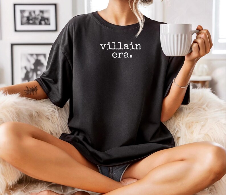 May include: Black t-shirt with the text "villain era." in white. The shirt is worn by a person holding a white mug. The person is sitting on a fluffy, white surface.