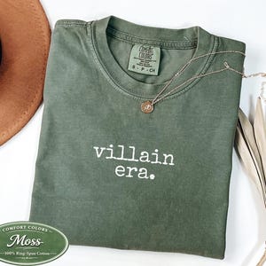 Moss Comfort Colors 1717 tee with villain era in white Special Elite typewriter font. Folded flat lay with brown cowboy hat, gold necklace, and dried palm. Earthy dark humor unhinged graphic tee.