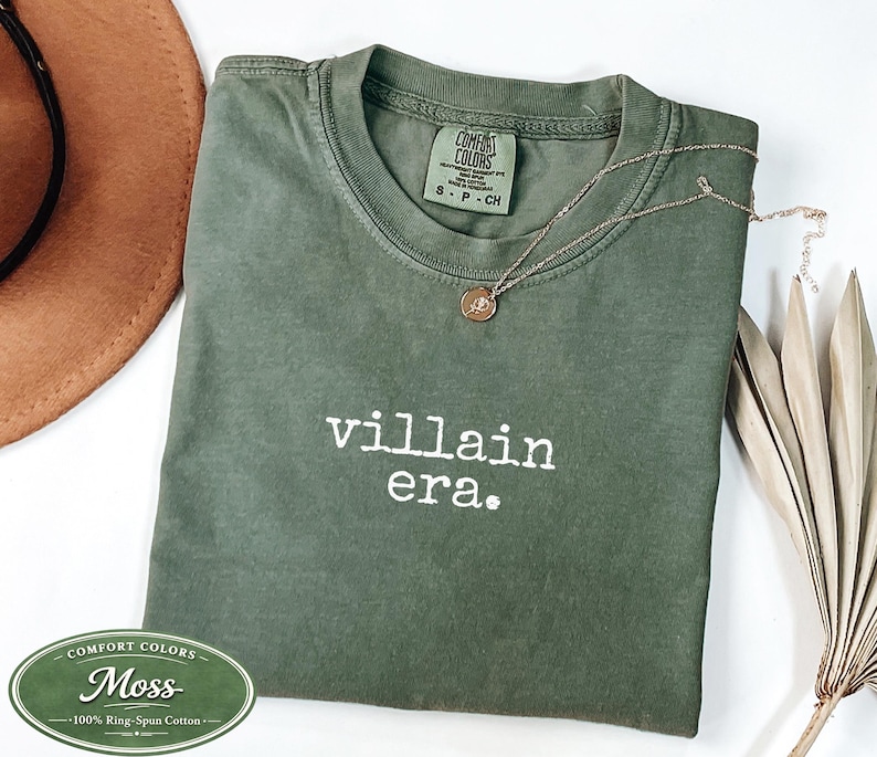 Moss Comfort Colors 1717 tee with villain era in white Special Elite typewriter font. Folded flat lay with brown cowboy hat, gold necklace, and dried palm. Earthy dark humor unhinged graphic tee.