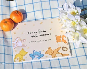 Può includere: Un libro intitolato "Dorky Buddies" con illustrazioni di animali dei cartoni animati, tra cui una volpe, un gatto e un orso, poggia su una superficie a scacchi blu e bianca. Sono presenti anche due arance e fiori bianchi.