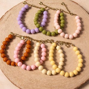 Op de afbeelding: Een verzameling armbanden met kralen in verschillende kleuren, waaronder paars, groen, roze, oranje, crème en geel. Elke armband heeft een ketting en wordt gepresenteerd op een houten oppervlak. De armbanden zijn gemaakt van ronde kralen.