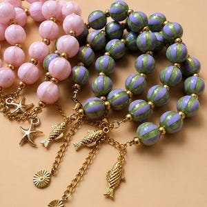 Op de afbeelding: Close-up van ketting met kralen met gouden accenten en bedels. Eén ketting heeft roze kralen, terwijl een andere groen en paars gestreepte kralen heeft. Bedels zijn onder meer zeesterren, vissen en zonnestraalontwerpen.