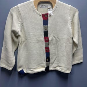May include: Cream-colored cardigan sweater with a round neck and long sleeves. The front features a vertical stripe of colorful squares in red, blue, black, and gray, fastened with wooden buttons. The brand name "ORVIS" is visible on the label.