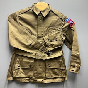 May include: A tan military-style jacket with multiple pockets, a belt, and a collar. The jacket has a patch on the upper arm with the word "AIRBORNE" and a symbol. The jacket is hanging on a wooden hanger.