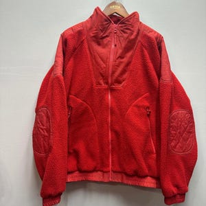 May include: A vibrant red fleece jacket with a high collar and a full-length zipper. The jacket features quilted patches on the elbows and shoulders, and two zippered pockets. The Orvis brand logo is visible on the hanger.