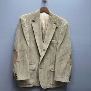 May include: A tan corduroy blazer with elbow patches. The blazer has a notched lapel, two front pockets, and a single-button closure. The sleeves have three buttons at the cuff. The blazer is hanging on a wooden hanger against a blue wall.