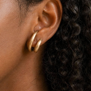 Peut inclure: Gros plan sur une oreille portant deux créoles dorées. Les boucles d'oreilles ont un design simple et arrondi. La personne a la peau foncée et des cheveux noirs bouclés. L'arrière-plan est de couleur claire et neutre.