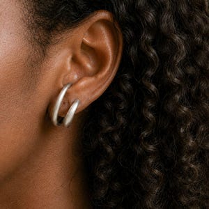 Peut inclure: Gros plan sur une oreille portant des créoles argentées. Les boucles d'oreilles sont de forme arrondie et simple. La personne a des cheveux bouclés foncés.