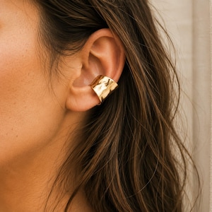 May include: A gold-colored ear cuff with a textured, hammered finish. The cuff wraps around the ear, creating a modern and stylish look. The jewelry piece is shown on a person with long, brown hair.