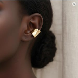 Peut inclure: Un ear cuff doré est porté sur l'oreille. Le bijou a une surface lisse et réfléchissante et un design géométrique. Le ear cuff est un bijou moderne.
