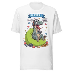 May include: White t-shirt featuring a cartoon dinosaur wearing headphones and playing a video game. The dinosaur is sitting on a green beanbag chair. Text above reads "PLAYER 1" with speech bubbles saying "BLAST!" and "NOOO!"