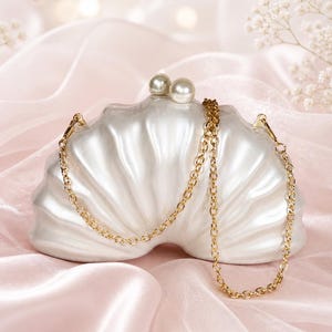 Peut inclure: Une pochette en forme de coquillage blanc nacré avec une bandoulière en chaîne métallique dorée. Le haut du sac présente deux fermoirs ronds semblables à des perles. La surface de la pochette a une finition lisse et brillante avec des stries.