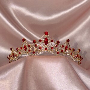 Op de afbeelding: Een goudkleurige metalen tiara met een ingewikkeld filigraanontwerp, versierd met talloze kleine heldere kristallen en diverse opvallende marquise- en rondgeslepen rode stenen in een symmetrisch patroon over de boogvormige kroon.