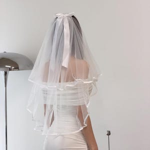 May include: A white, two-tiered bridal veil with a satin ribbon bow at the crown. The veil is made of a sheer, delicate fabric and is paired with a white wedding dress. The dress has a ruched bodice and a fitted skirt.