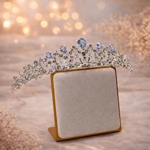 May include: A silver-toned tiara featuring intricate metal leaf and vine patterns adorned with small clear crystals and five prominent, light blue, pear-shaped crystals arranged along the top edge, resting on a square display stand.
