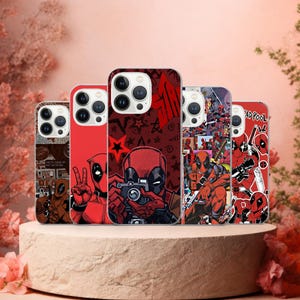 Puede incluir: Cinco fundas para teléfono con diseños de Deadpool en rojo y negro. Las fundas muestran varias imágenes del personaje, algunas con texto. Se exhiben sobre un pedestal marrón claro, con un fondo rosa suave.