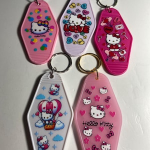 May include: Six Hello Kitty keyrings in pink, white, and lilac. Each keyring features a different design with the iconic character, hearts, bows, and the words "Hello Kitty" or "Love". The keyrings have metal rings.