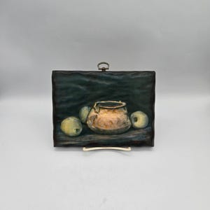May include: A rectangular wall plaque featuring a painted still life of a metallic copper-colored bowl centered between three pale green apples against a dark, textured background. The plaque has a wavy edge and a small metal hanging loop at the top.