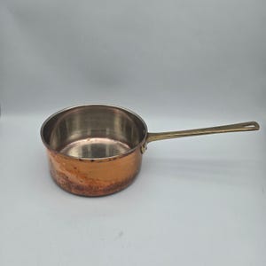 May include: A round copper saucepan with a polished silver-toned interior and a long, slender brass handle. The exterior of the pan features a warm, metallic copper finish with visible signs of natural patina and wear along the base.