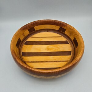 May include: A round wooden bowl featuring a segmented design with alternating light tan and dark brown wood strips. The interior base displays horizontal stripes of contrasting wood tones, while the rim showcases a repeating block pattern.