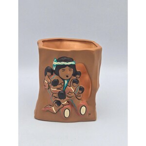 May include: A terracotta-colored ceramic vase with a rectangular shape. The vase features a relief design of a Native American figure surrounded by children. The figures are painted with black, blue, and white accents. The vase has a textured, bark-like exterior.