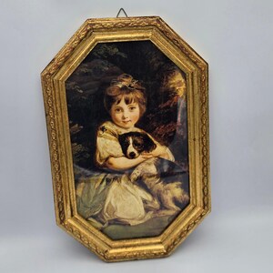 May include: An octagonal framed print featuring a portrait of a child with brown hair wearing a white blouse, holding a black and white dog. The frame is ornate, metallic gold with a textured, repeating pattern along the edges and a small metal hook.
