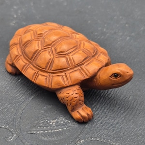 May include: A small, hand-carved wooden turtle figurine with a smooth, light brown finish. The sculpture features detailed, raised hexagonal patterns on the shell, textured scales on the legs, and a defined head with a small, dark, inlaid eye.