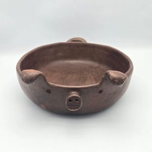 May include: A round, dark brown clay bowl shaped like a pig. The bowl features two small ears on the sides, a protruding snout with two nostrils in the center, and two small indentations for eyes. The surface has a matte, textured finish.