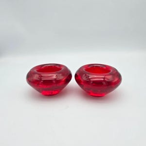 May include: Two circular, bowl-shaped candle holders made of translucent red glass. Each piece features a smooth, rounded exterior with a wide, recessed center opening designed to hold a small candle, showcasing a vibrant, deep red hue.