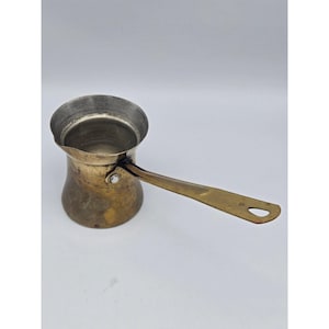 May include: A brass Turkish coffee pot with a wide, flared rim and a long, flat handle featuring a small hole at the end. The body has a rounded base and a metallic finish with visible signs of wear and oxidation, showing a warm, golden-brown tone.