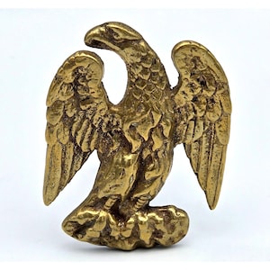 May include: A metallic gold-toned eagle emblem featuring a textured finish with the bird depicted in profile, facing left with wings partially spread and talons resting on a base. The surface shows a weathered, antique-style patina throughout.